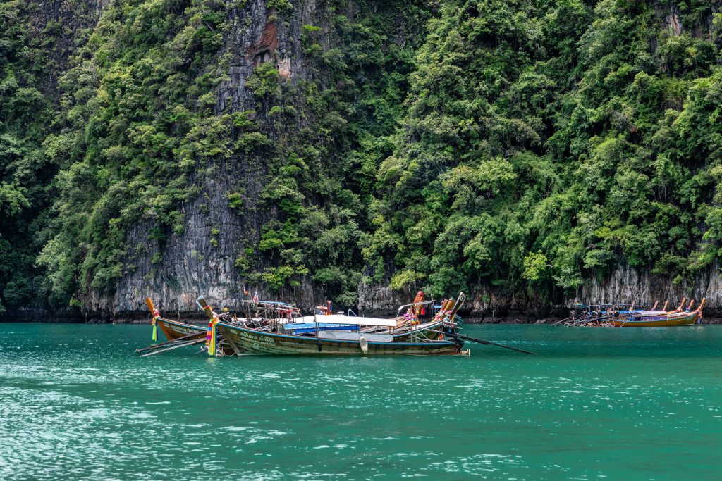 Traditional wooden Thai longtail boats floating on calm turquoise water against a backdrop of steep limestone cliffs covered in lush tropical greenery.