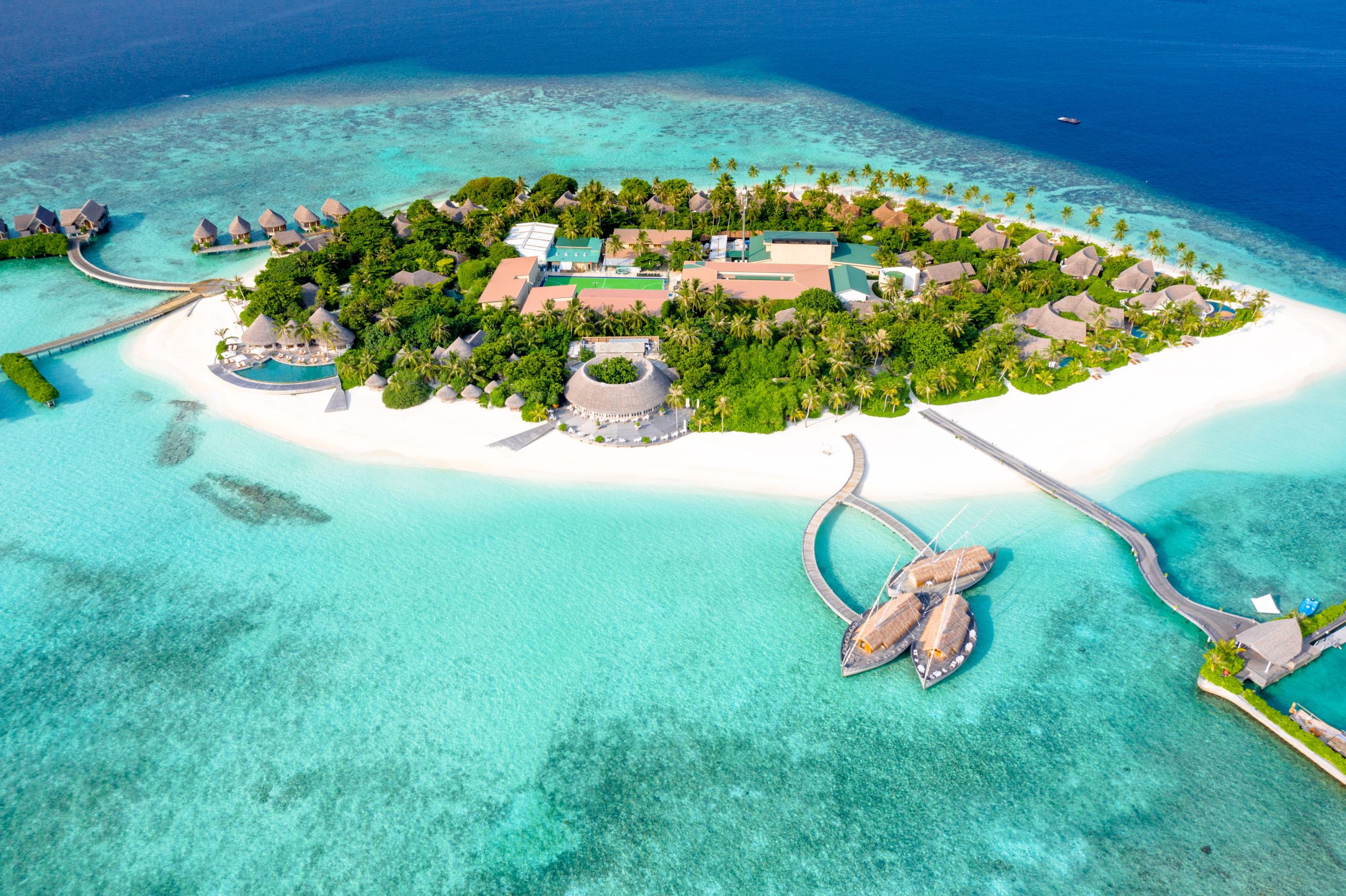 Breathtaking drone shot of a tropical island resort in the Maldives featuring lush green palm trees, pristine white sand beaches, and turquoise coral waters surrounded by deep blue ocean.