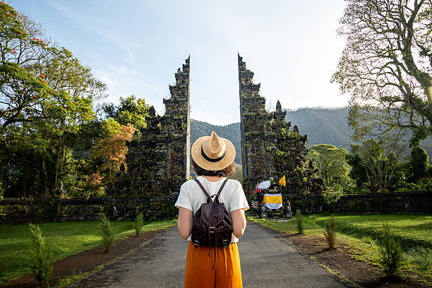 A female traveler with a backpack and straw hat standing on a pathway facing the majestic traditional Balinese split gates (Candi Bentar) at Handara Golf & Resort in Bali, Indonesia.