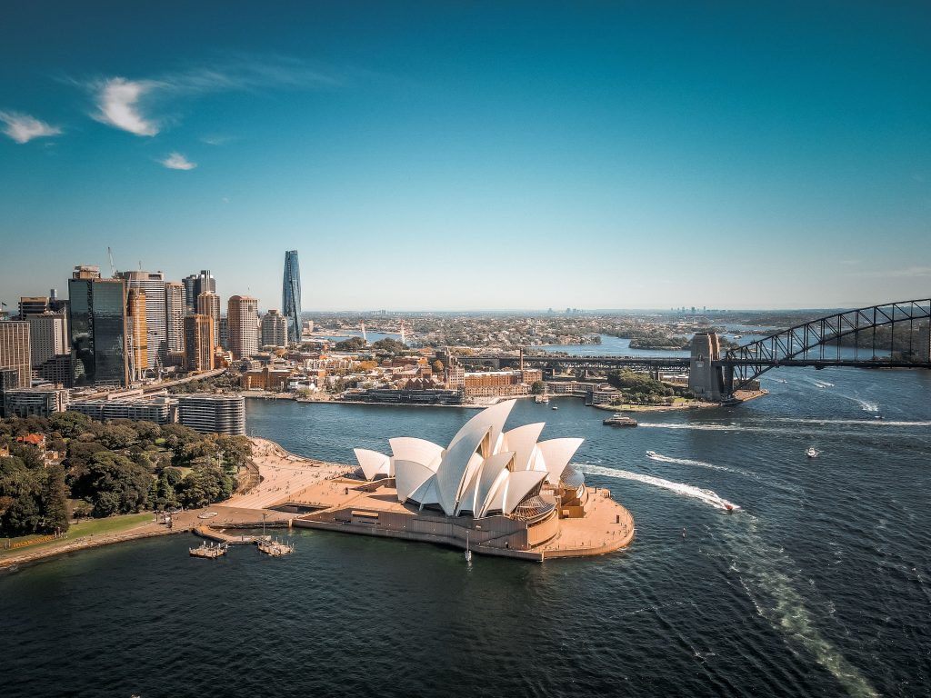 High-angle aerial view of the iconic Sydney Opera House situated on Sydney Harbour, with the Sydney Harbour Bridge and city skyline in the background under a clear blue sky.