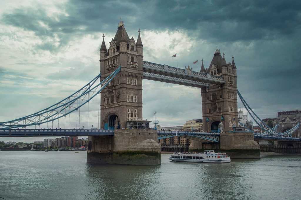 The majestic Tower Bridge with its iconic stone towers and suspension spans crossing the River Thames in London under a dramatic cloudy sky, with a tourist boat passing underneath.