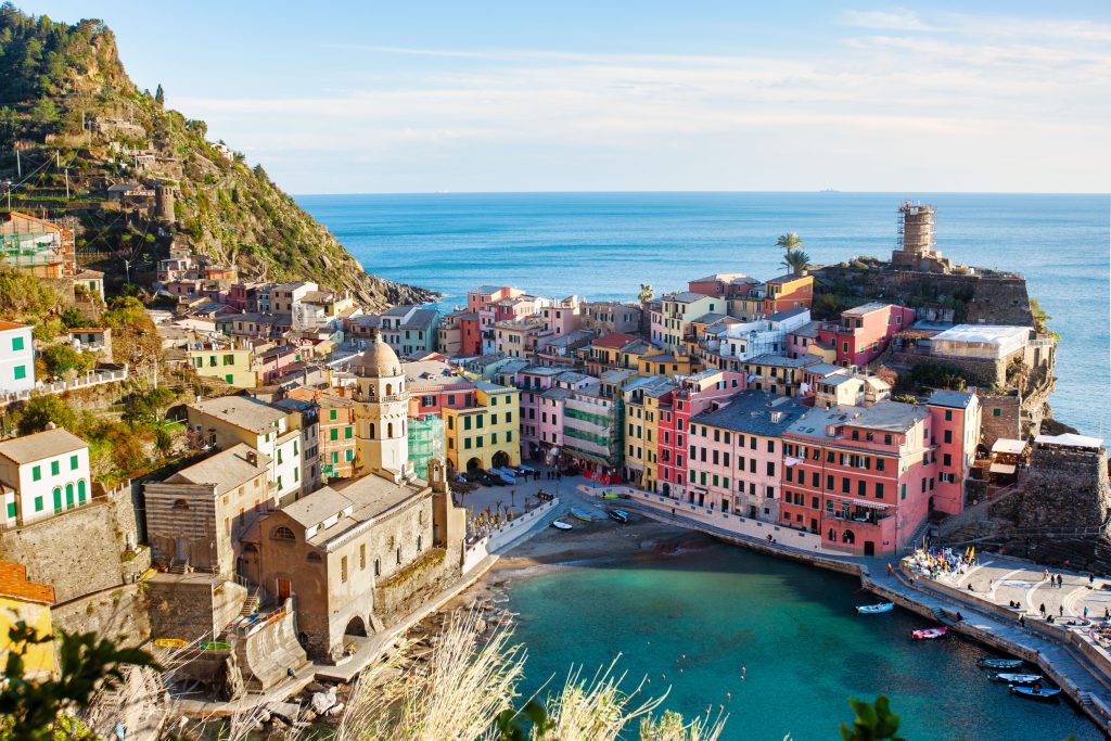 A stunning elevated view of the colorful historic coastal village of Vernazza in Cinque Terre, Italy, featuring pastel buildings, a small harbor with turquoise water, and rugged green cliffs overlooking the sea.