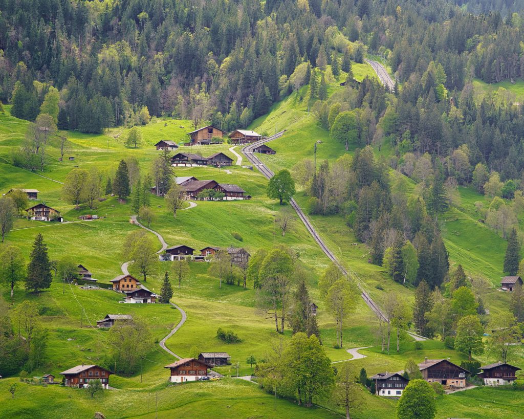 A charming Swiss alpine village with traditional wooden chalets scattered across lush green sloping hills, connected by winding roads and a mountain funicular railway track running up through the landscape in Switzerland.