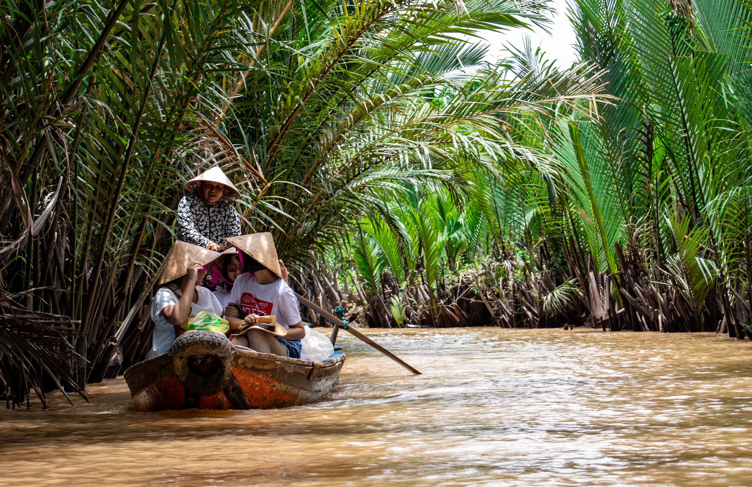 Tourists wearing traditional conical hats riding a wooden boat steered by a local guide through a narrow river channel lined with lush coconut palms in Vietnam.